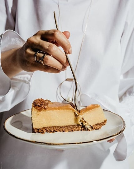 Main tenant un verre de glaçage au-dessus d'une tranche de cheesecake garnie sur assiette blanche