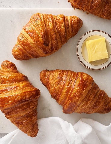 Trois croissants dorés et feuilletés disposés sur papier blanc avec un petit bol de beurre frais