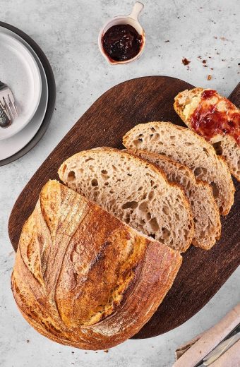 Pain artisanal tranché sur planche en bois avec confiture et accessoires de table, vue de dessus