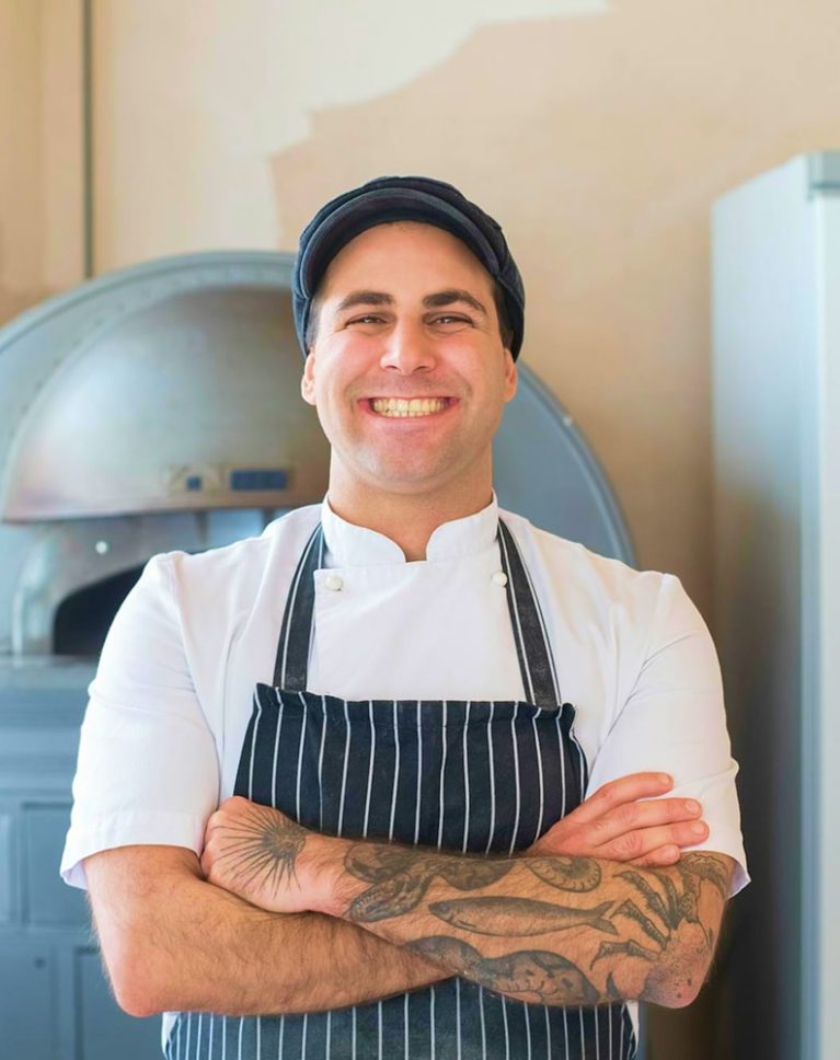 Boulanger souriant en tenue professionnelle devant un four à pain traditionnel en pierre