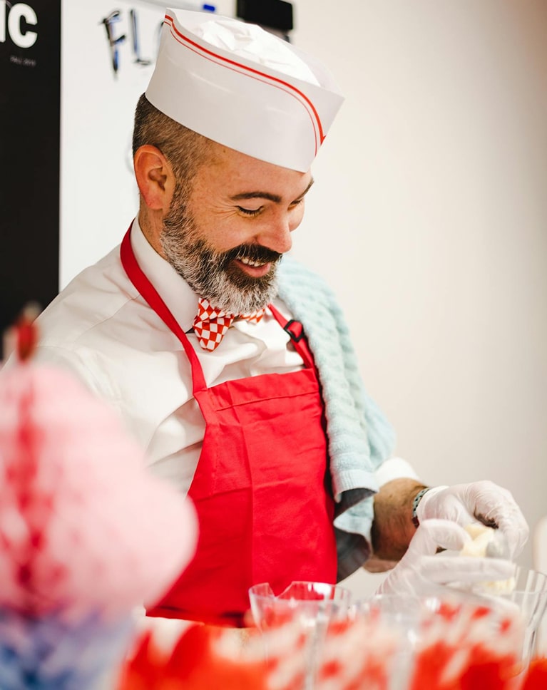 Chef pâtissier portant un tablier rouge et un chapeau blanc avec nœud papillon à carreaux rouges et blancs, préparant des