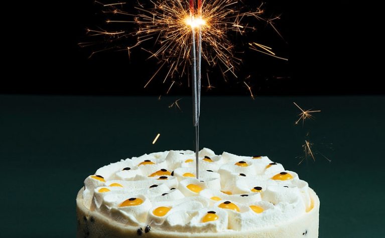 Gâteau blanc décoré de fruits confits avec un cierge magique étincelant sur fond bleu-vert