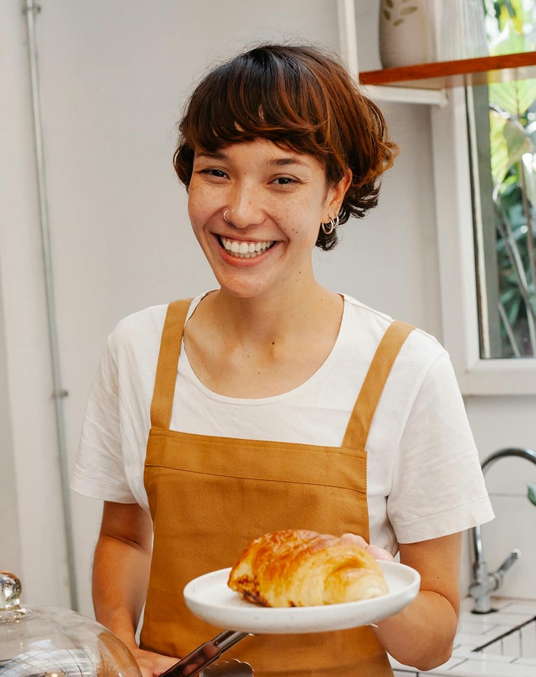 Femme souriante en tablier marron tenant une assiette avec un croissant doré dans une cuisine lumineuse