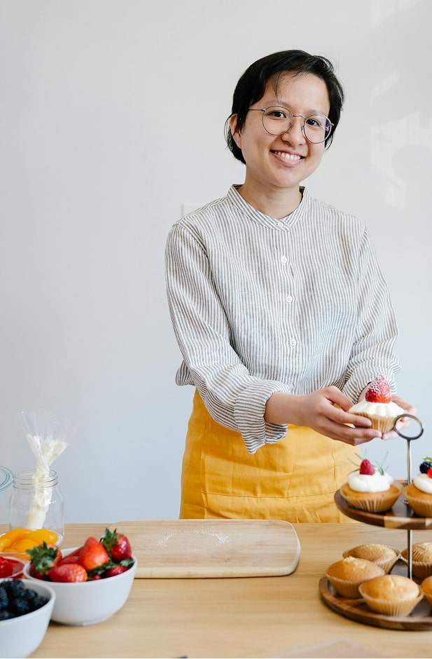 Boulangère souriante en chemise grise et tablier jaune présentant un cupcake garni de fraise, avec pains et pâtisseries sur