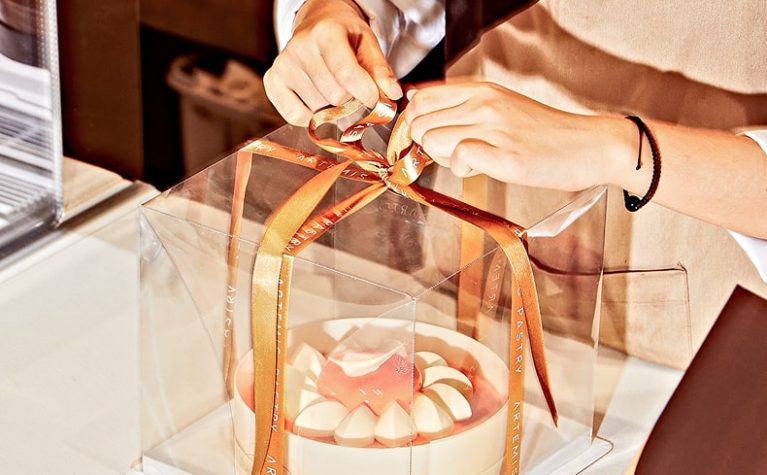 Personne en tablier attachant un ruban doré autour d'une boîte transparente contenant un gâteau blanc avec décoration