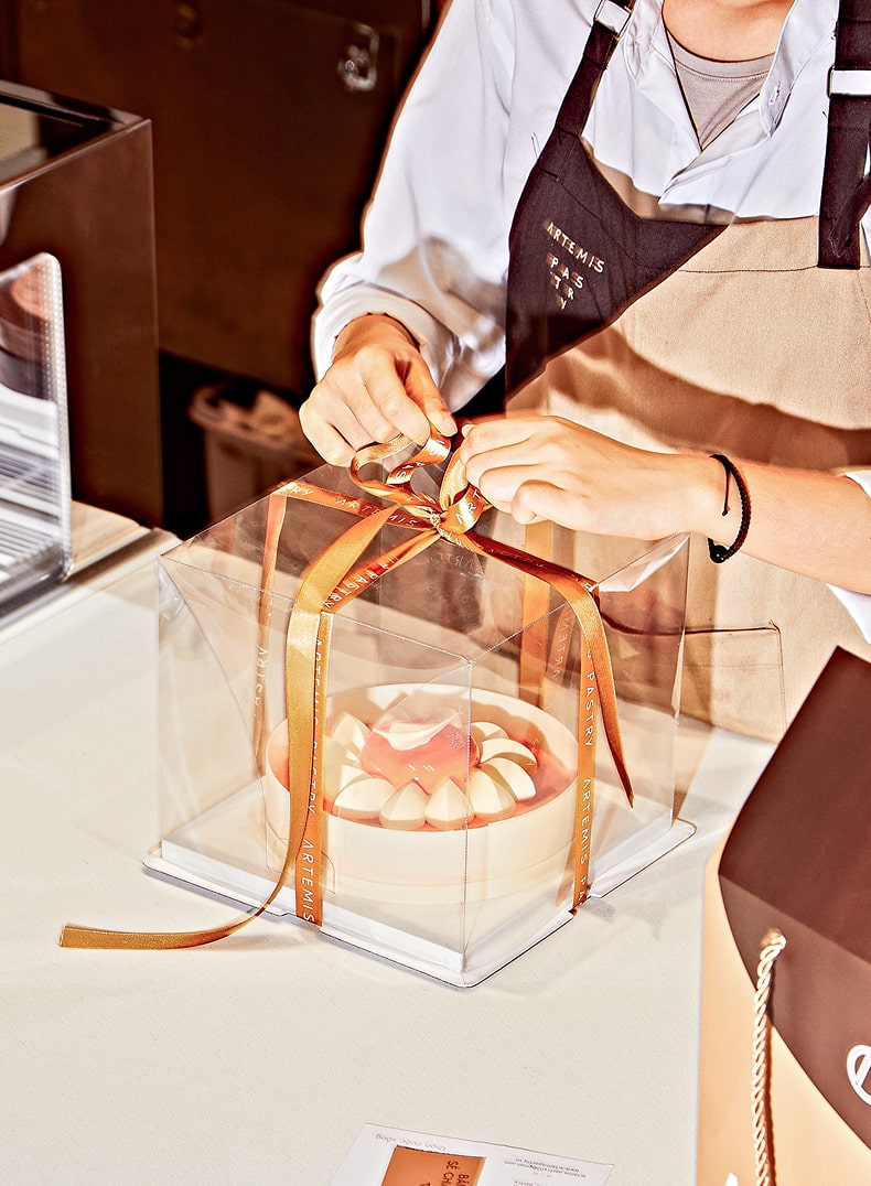Personne en tablier attachant un ruban doré autour d'une boîte transparente contenant un gâteau blanc avec décoration