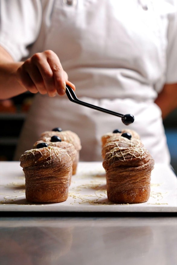 Pâtissier décorisant des muffins individuels avec un glaçage noir à l'aide d'une cuillère, sur un plateau blanc