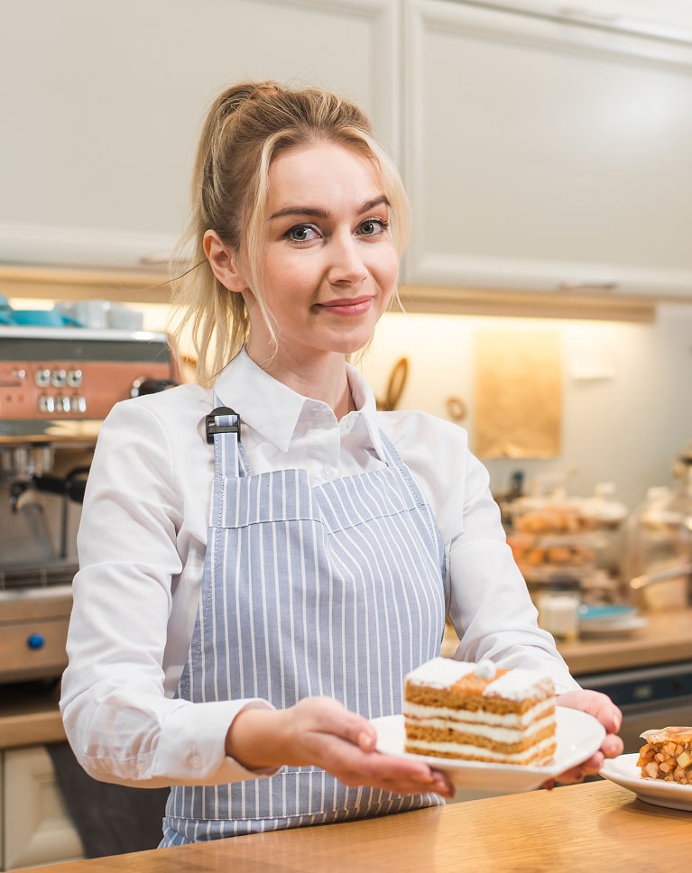 Jeune femme boulangère en tablier bleu à rayures présentant une part de gâteau en couches dans une cuisine professionnelle
