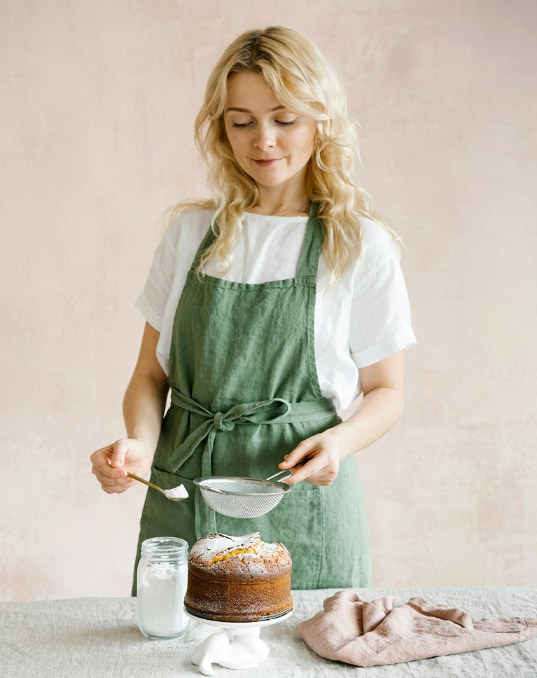 Femme blonde en tablier vert saupoudrant du sucre glace sur un gâteau avec un tamis