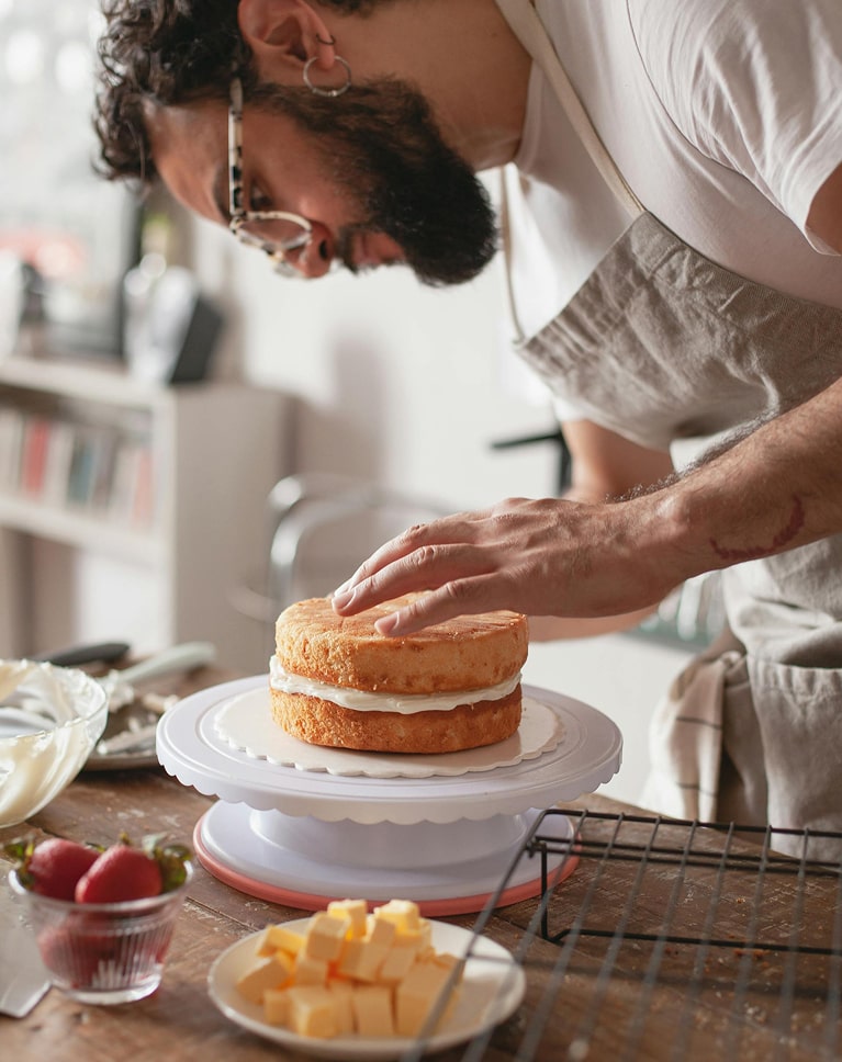 Boulanger en train de décorer un gâteau à deux étages avec glaçage blanc sur un plateau tournant