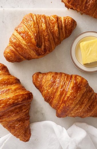Trois croissants dorés et beurrés disposés sur papier blanc avec un petit bol de beurre frais
