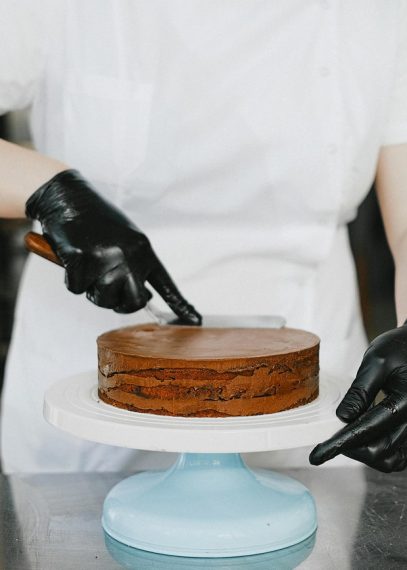 Pâtissier en gants noirs et tenue blanche garnissant un gâteau chocolat en étages sur un présentoir bleu ciel