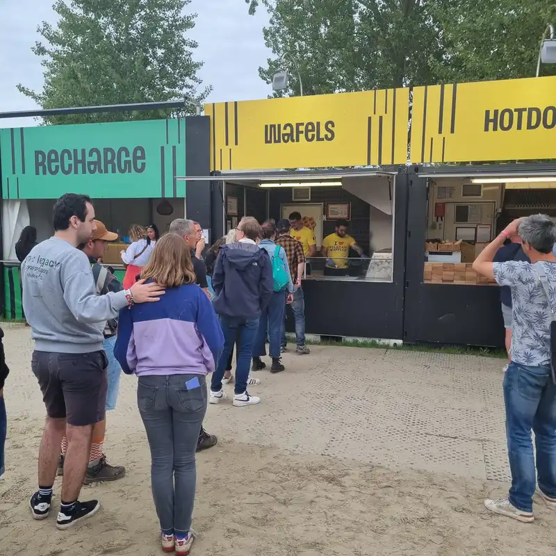 File d'attente de clients devant des food trucks Recharge, Wafels et Hotdog dans un parc avec arbres