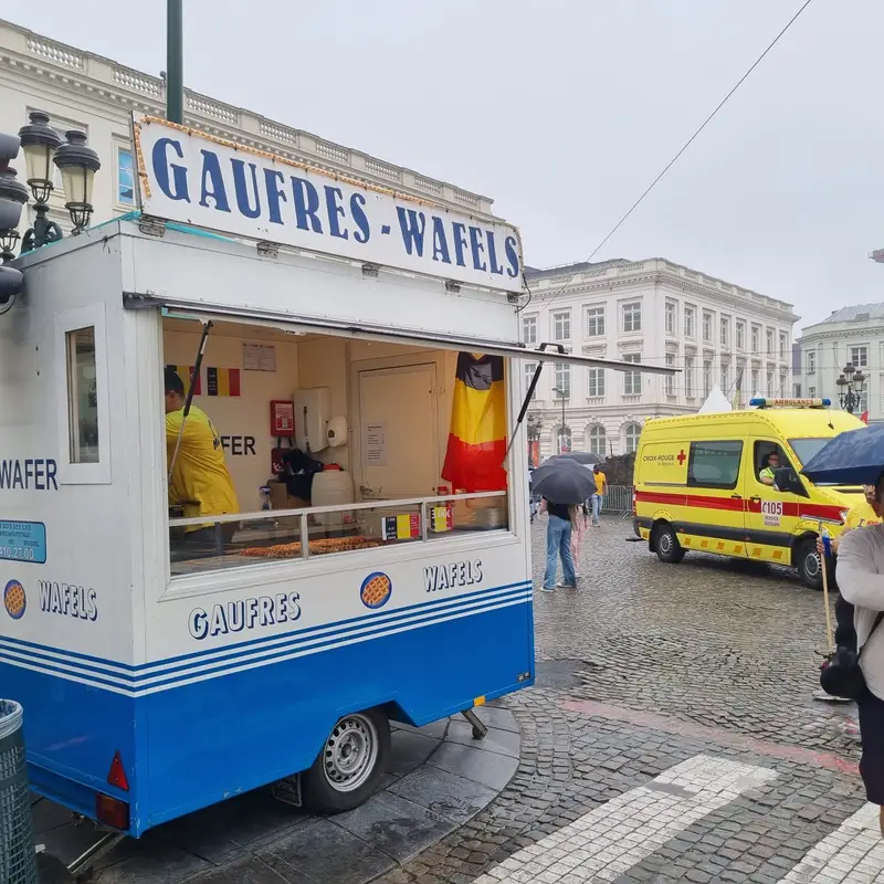 Food truck Gaufres-Wafels blanc et bleu avec vendeur en tenue jaune-rouge sur une place pavée en Belgique, ambulance en