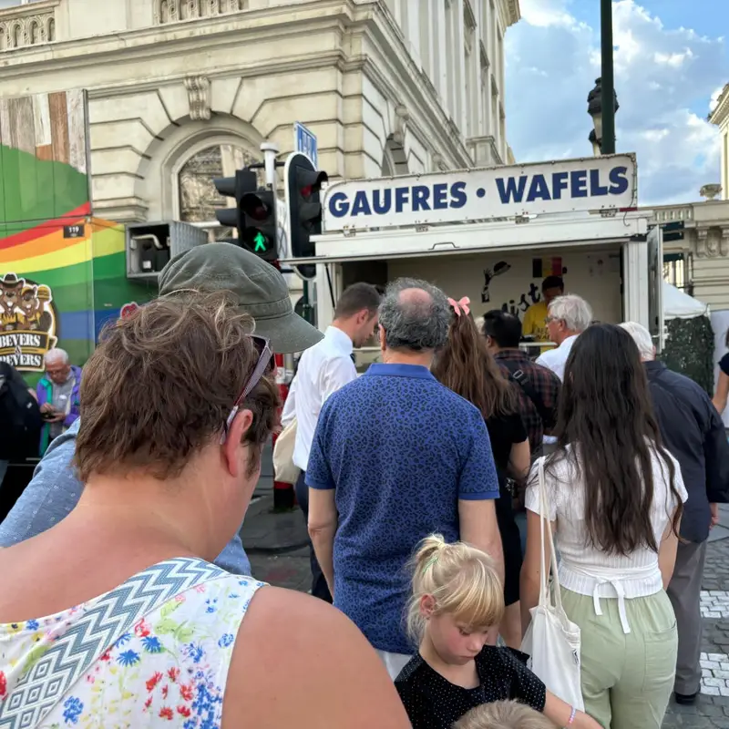 File d'attente de clients devant un food truck Gaufres Wafels à un événement public avec drapeaux arc-en-ciel et bâtiments
