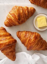 Trois croissants dorés et feuilletés disposés sur papier blanc avec un petit bol de beurre frais