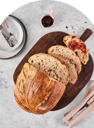 Pain artisanal tranché sur planche à découper en bois avec couteau, assiette et confiture sur surface grise