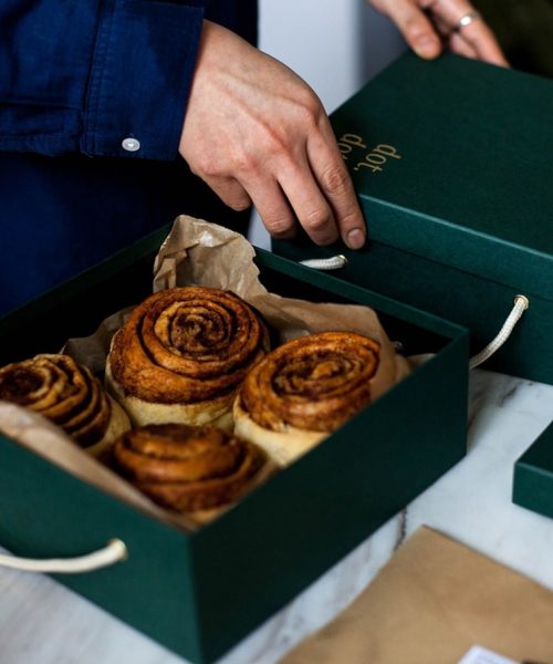 Homme en chemise bleue ouvrant une boîte verte contenant quatre spirales de pain à la cannelle dorées sur papier kraft