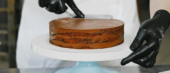 Pâtissier en gants noirs présentant un gâteau éponge multicouche sur un présentoir blanc
